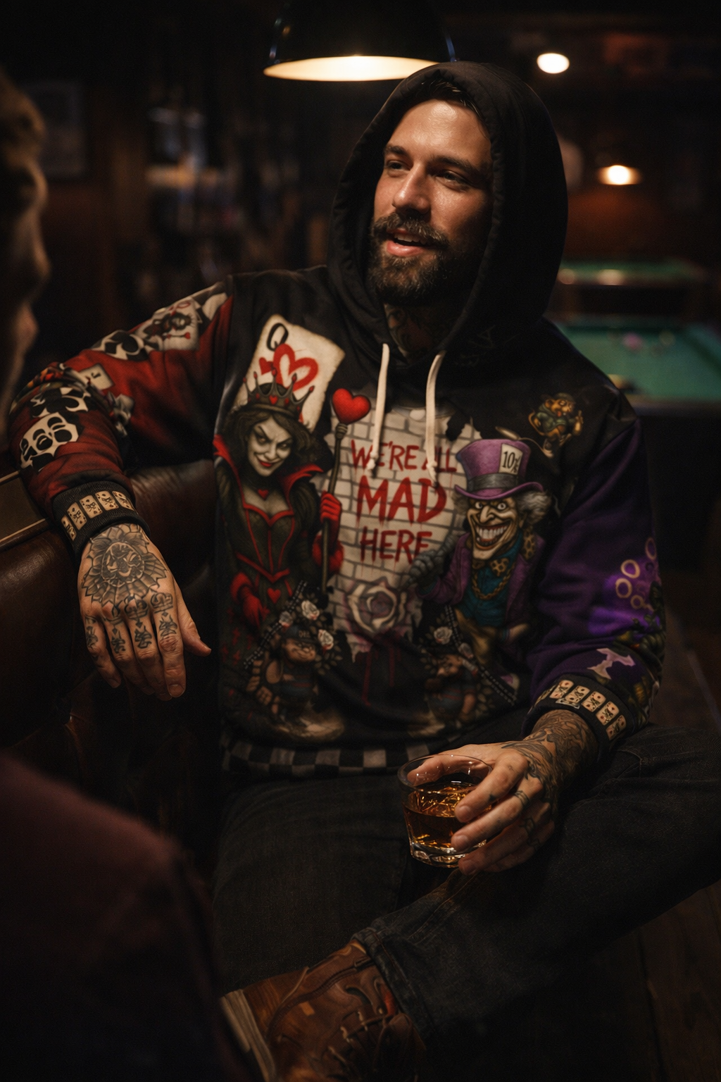 Man sitting in a bar wearing a hoodie with various designs, holding a drink.